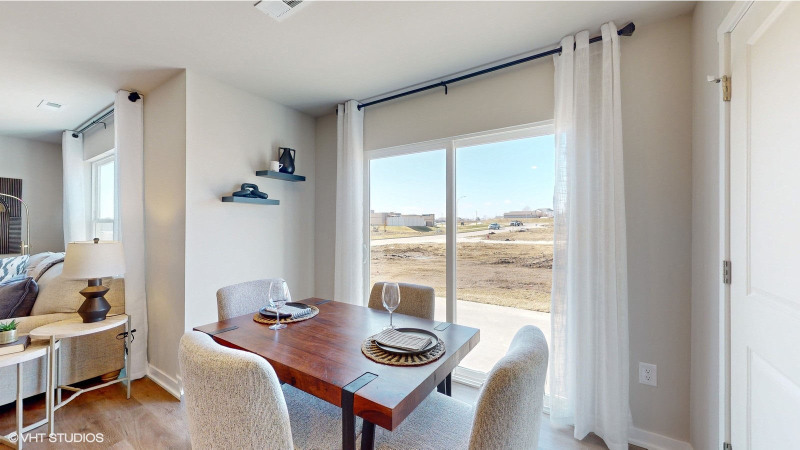 A cozy dining nook featuring a modern wooden table set for two with upholstered chairs, situated beside large sliding glass doors that allow for abundant natural light and views of the open landscape beyond.