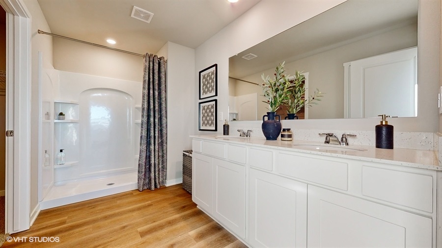 Modern bathroom with wood flooring, a white double sink vanity, and a large mirror. A shower with a patterned curtain is on the left. Decorative plants and framed art add elegance.