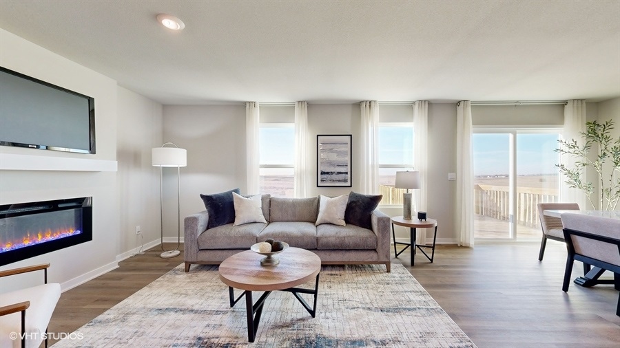 Modern living room with neutral tones featuring a gray sofa, cushions, round wooden coffee table, abstract art, and large windows offering scenic views. A sleek fireplace adds warmth, while a soft rug enhances coziness.