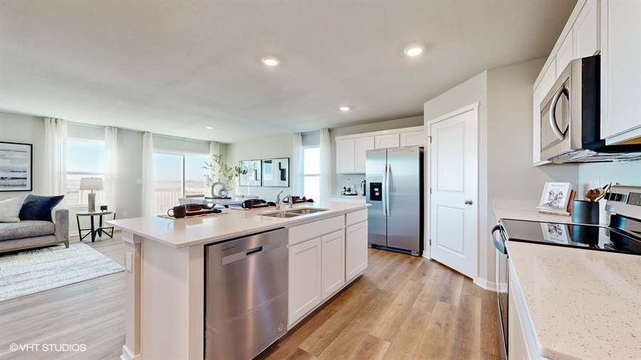 Modern kitchen with white cabinets, stainless steel appliances, and a center island with sink. Connected living area with sofa and large windows. Bright, airy ambiance.