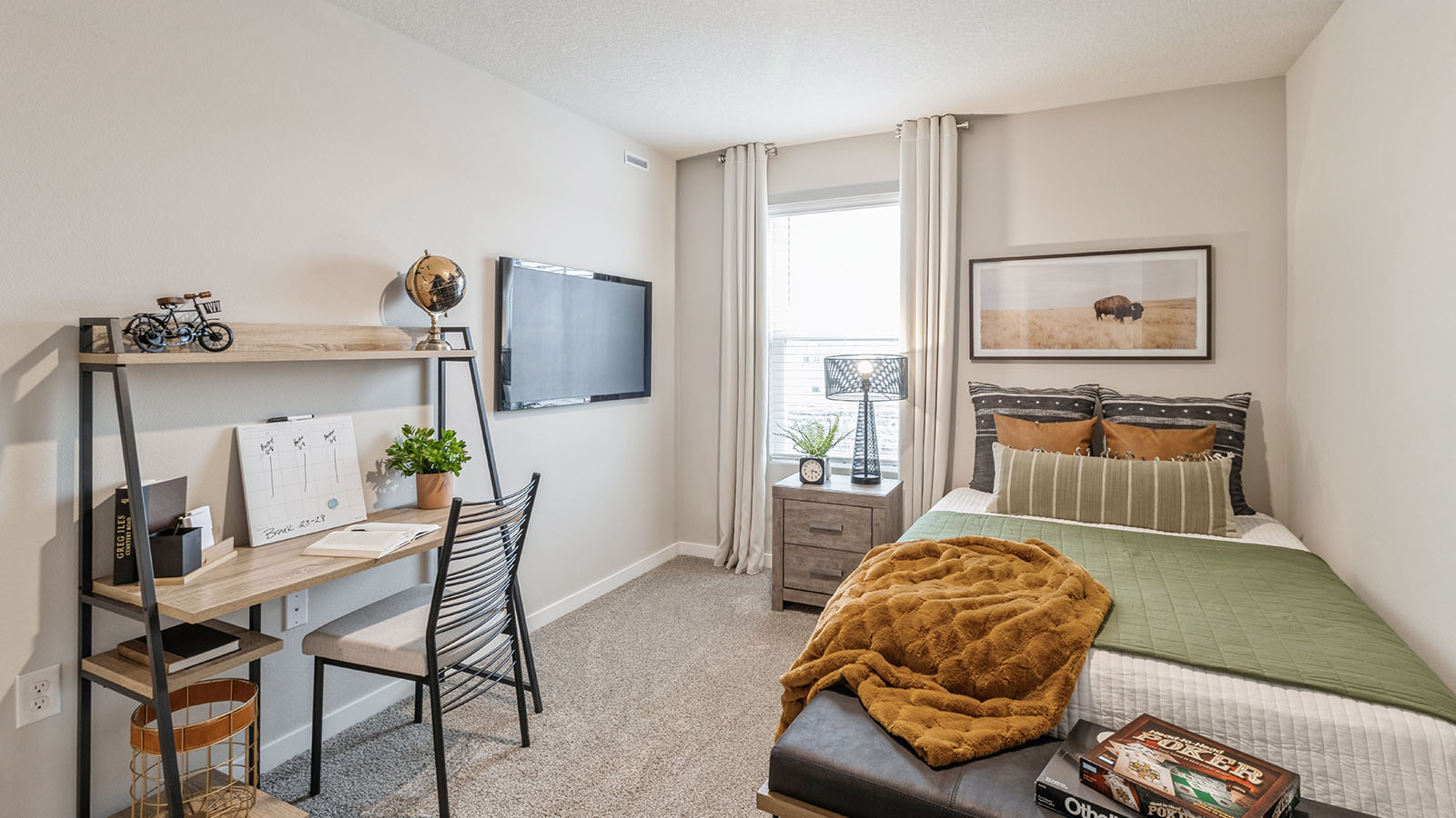 A cozy bedroom with a twin bed, earth-toned bedding, a small desk and shelving unit, a wall-mounted TV, and a window with white curtains.