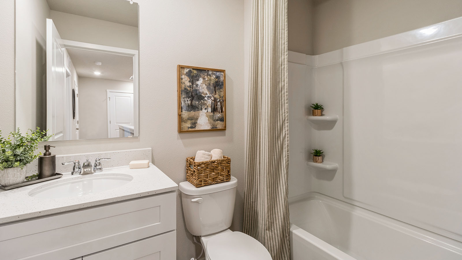 A neutral-toned bathroom featuring a white vanity, a bathtub with a shower curtain, small built-in shelves, and a decorative basket with towels on the toilet.