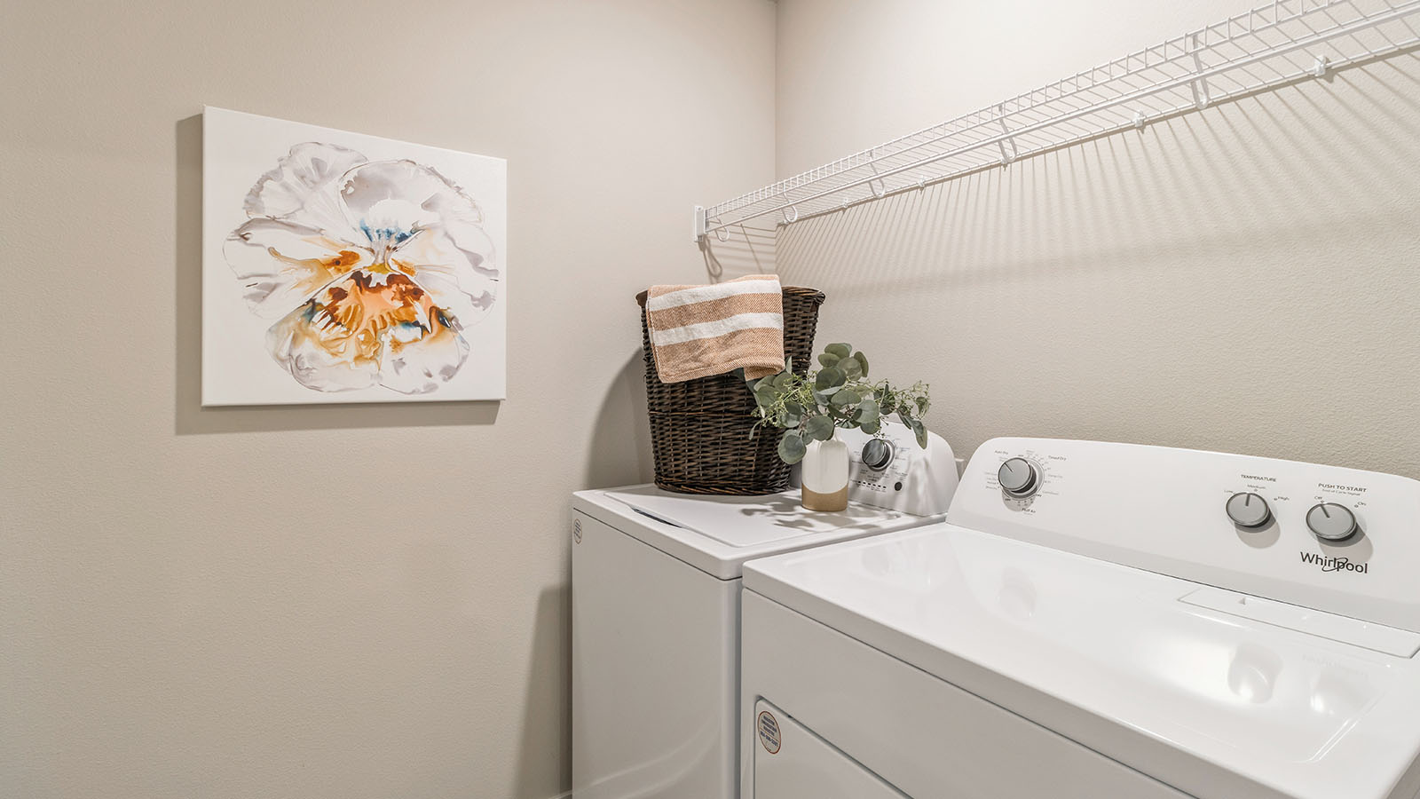 A compact laundry room with a top-loading washer and dryer, a wicker basket with folded towels, decorative greenery, and a floral wall art piece.