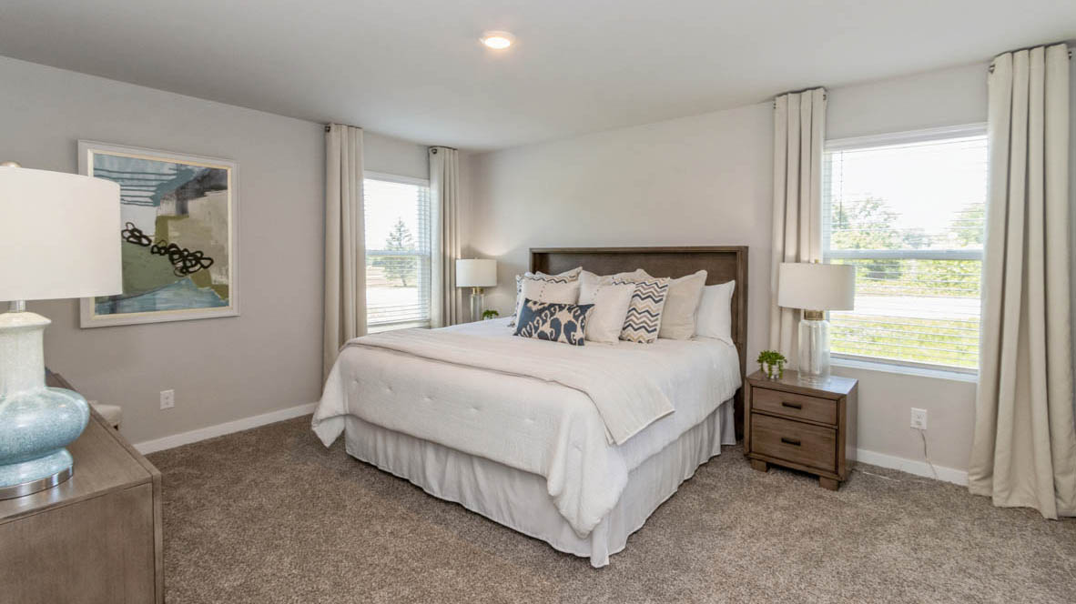 The Neuville's carpeted primary bedroom with a bed, two nightstands, and a dresser with natural light from windows on two walls.