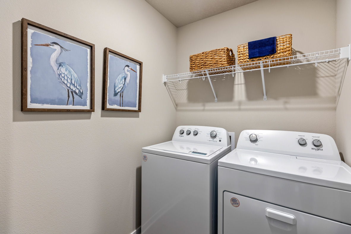 Laundry room in the Roland with a full-sized washer and dryer, shelf for storage, and art prints on the wall.