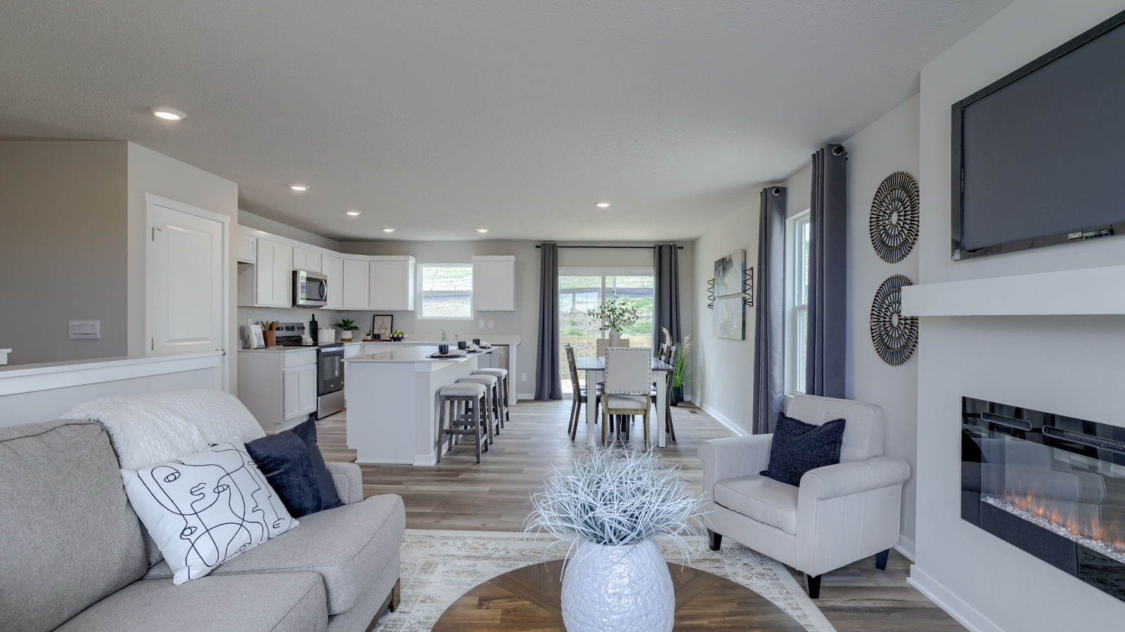 Spacious living room with brown flooring, white walls, a black fireplace and windows and kitchen