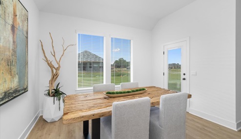 Dinning are featuring a large wooden table with gray chair. The room has two large windows allowing natural light, with white walls, and light wood floor.