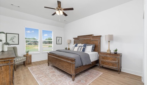 Master bedroom featuring a wooden bed frame with matching nightstands with a gray comforter and decorative pillows. The room has two large windows allowing natural light, with white walls, a light wood floor, and a ceiling fan.