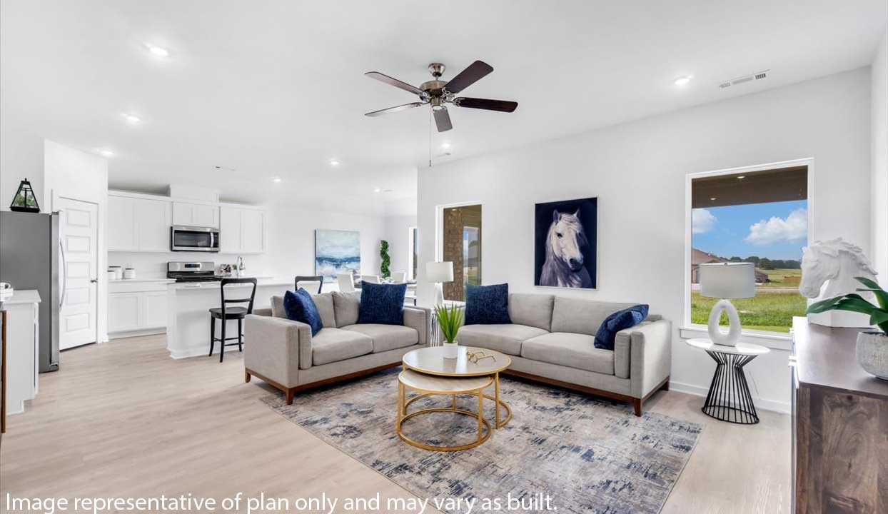 Open-concept living room with light gray sofas, blue accent pillows, a horse portrait, and large windows. Adjacent modern kitchen with white cabinetry and stainless steel appliances. Ceiling fan and round coffee table complete the cozy, airy space.