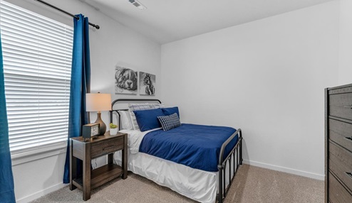 Cozy bedroom with a metal-framed bed featuring blue bedding and decorative pillows, a wooden nightstand with a lamp, and artwork of animals on the wall. The room has carpeted flooring, a window with blue curtains, and a dark wood dresser.