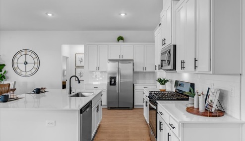 Modern kitchen with white cabinets, stainless steel appliances, and a sleek white quartz countertop. Features include a center island with a built-in sink, a gas stove, and a decorative clock on the wall. The space is accented with plants and minimalistic decor.