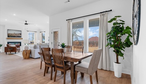 Open-concept dining and living area with a wooden dining table and chairs, large windows with beige curtains, and a vibrant potted plant. The living room features a cozy seating area, a fireplace with a wood mantel, and a mounted landscape painting.
