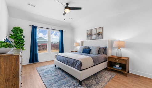 Bright and airy bedroom with a large upholstered bed featuring blue and neutral bedding, flanked by wooden nightstands with matching lamps. The room includes a vaulted ceiling with a ceiling fan, large windows with blue curtains, and a decorative rug. A potted plant and a wooden dresser add warmth and natural elements.