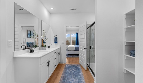 Modern bathroom with a double vanity featuring white countertops and black faucets, large mirrors, and decorative orchid accents. A glass-enclosed shower is visible, and the space connects to a bedroom with large windows. Built-in shelving adds functionality, and a blue patterned rug completes the design.