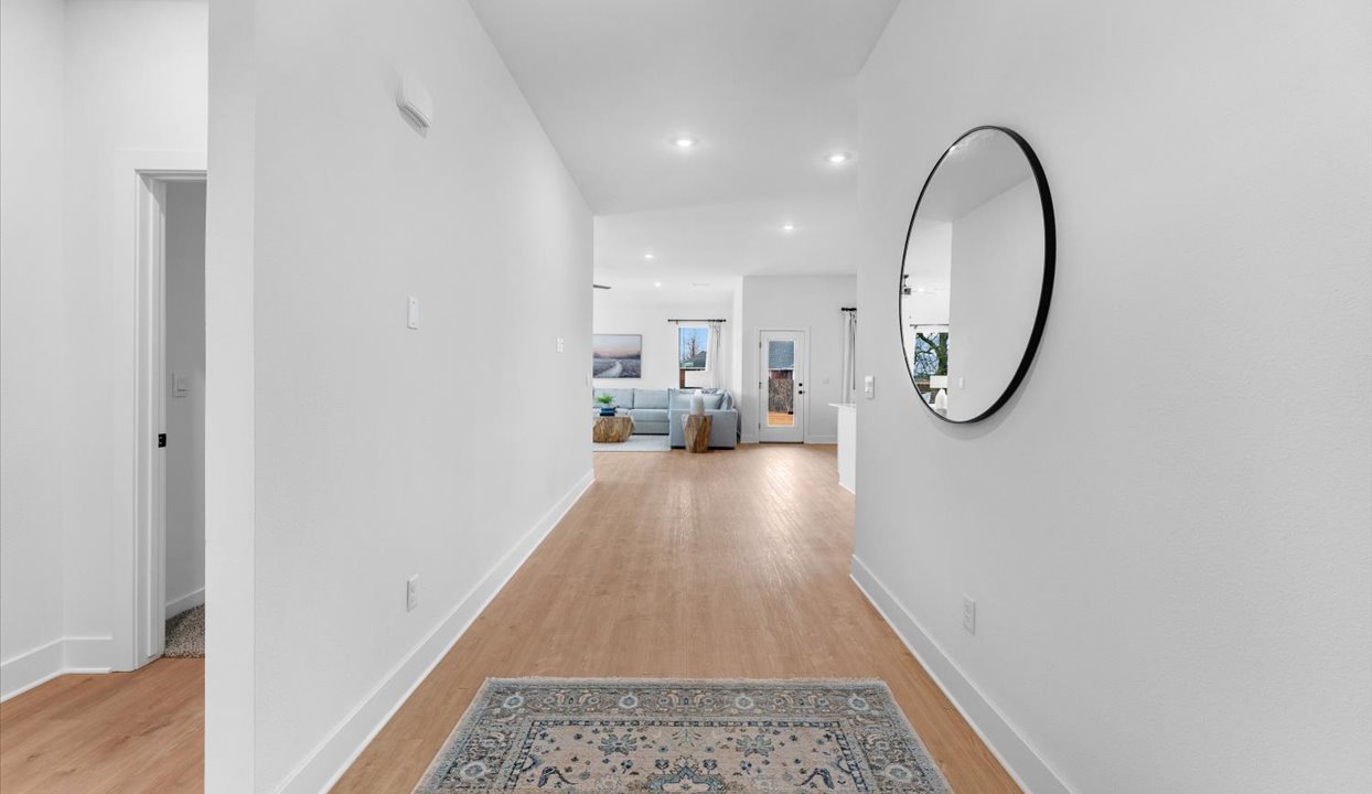 Interior hallway with white walls, a wood floor, and a decorative round mirror mounted on the wall, leading to a bright living area with large windows and modern furniture.