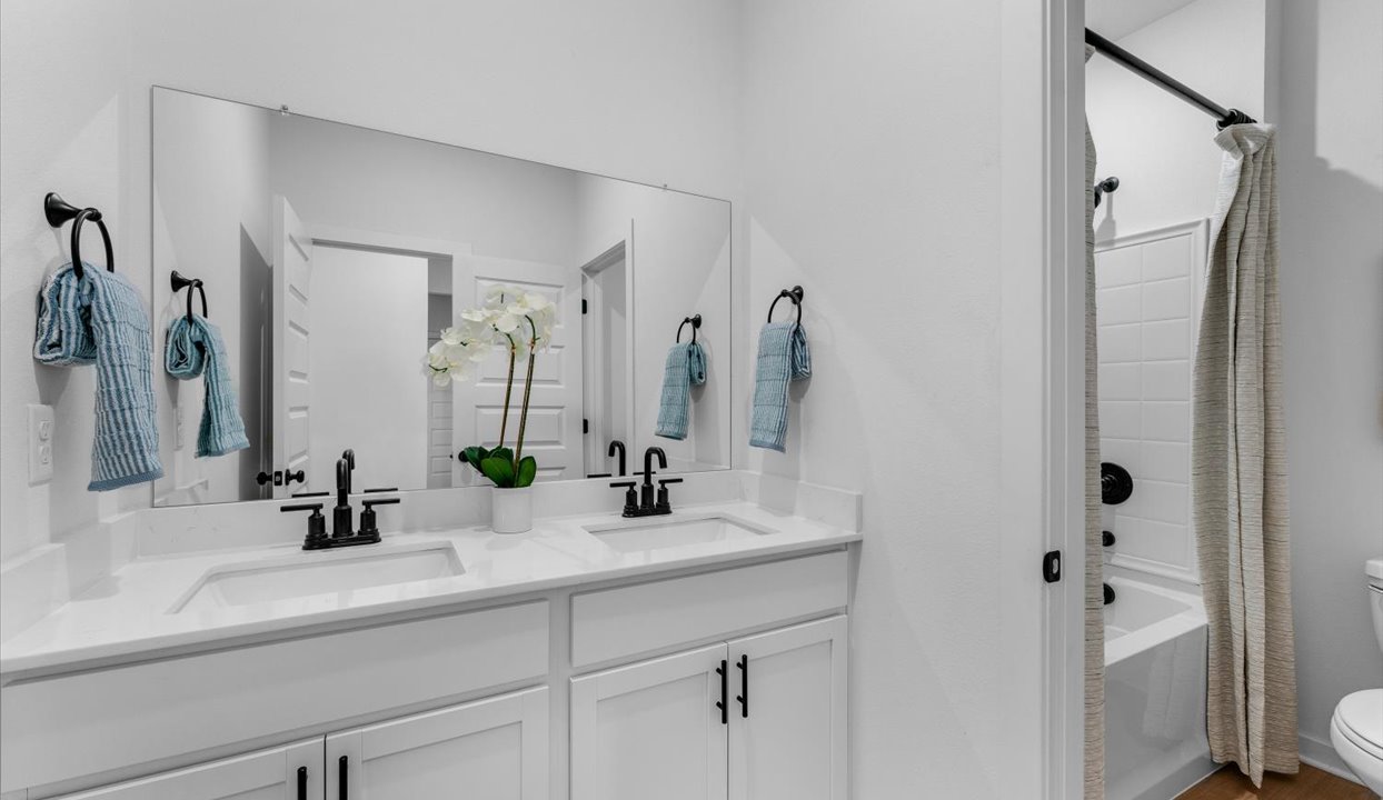 Modern bathroom featuring a double vanity with white countertops, black faucets, and a large mirror. Decor includes light blue hand towels and a white orchid plant. The room also includes a bathtub with a shower curtain and white tiled walls.