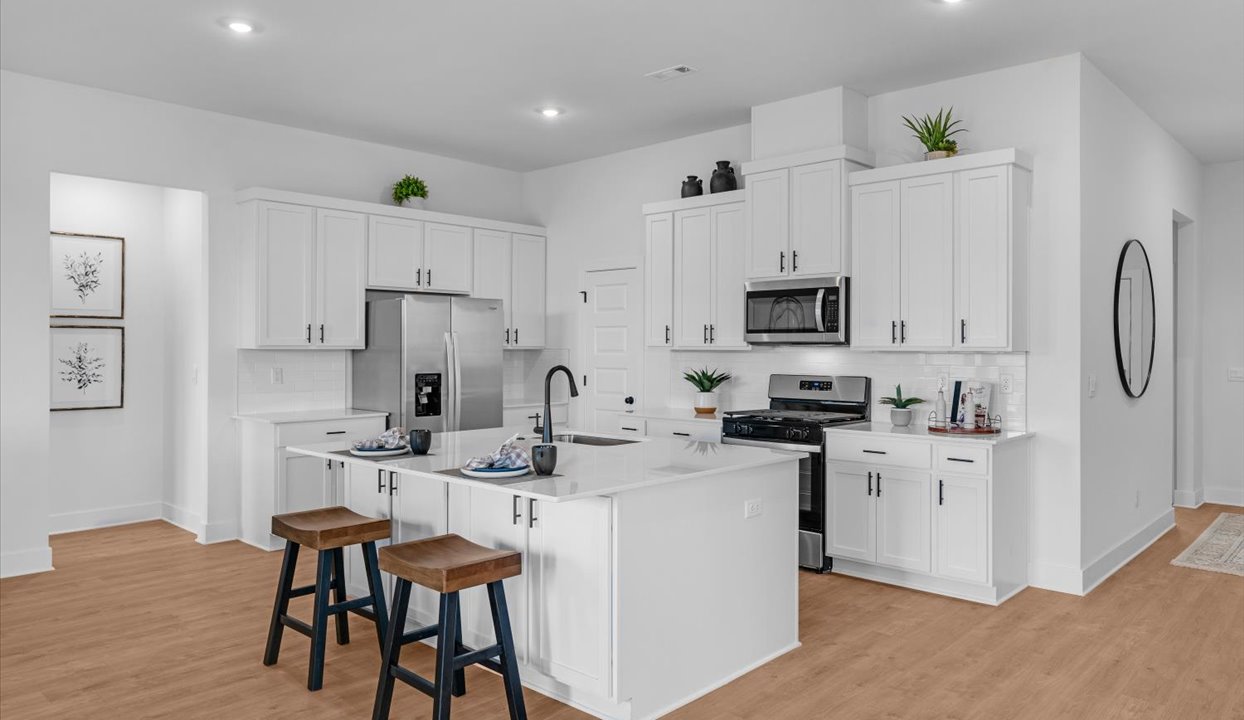 Bright, modern kitchen with white cabinetry, stainless steel appliances, and a large island featuring a built-in sink and seating for two with wooden stools. Accents include greenery, framed artwork, and decorative items for a welcoming touch.
