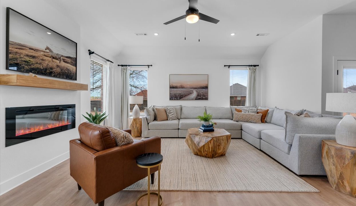 Spacious open-concept living area with a light gray sectional sofa, a wooden geometric coffee table, and a leather accent chair. The room features modern decor, a ceiling fan, and a view of the adjacent kitchen with white cabinetry and a center island.