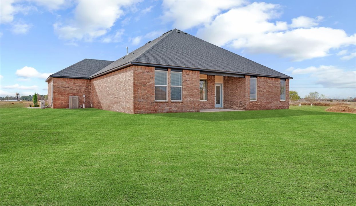 Covered outdoor patio with brick walls, large windows, and a concrete floor, overlooking a well-maintained green lawn and a scenic backyard view.