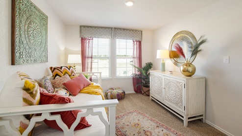 San Antonio Texas Express Homes DR Horton Applewood Model Home Jasmine floor plan 2182 square feet New Construction Homes secondary or guest bedroom with carpet flooring large windows for natural lighting sheer pink curtains with valence house plant twin sized day bed with white frame and comforter chevron faux fur throw pillows colorful throw pillows all sizes copper patina mandala mounted wall art table lamps small quilted ottoman artisan area rug white wooden chest of drawers with mandala patterned doors brass table vase with decorative feathers and mounted circular brass wall art