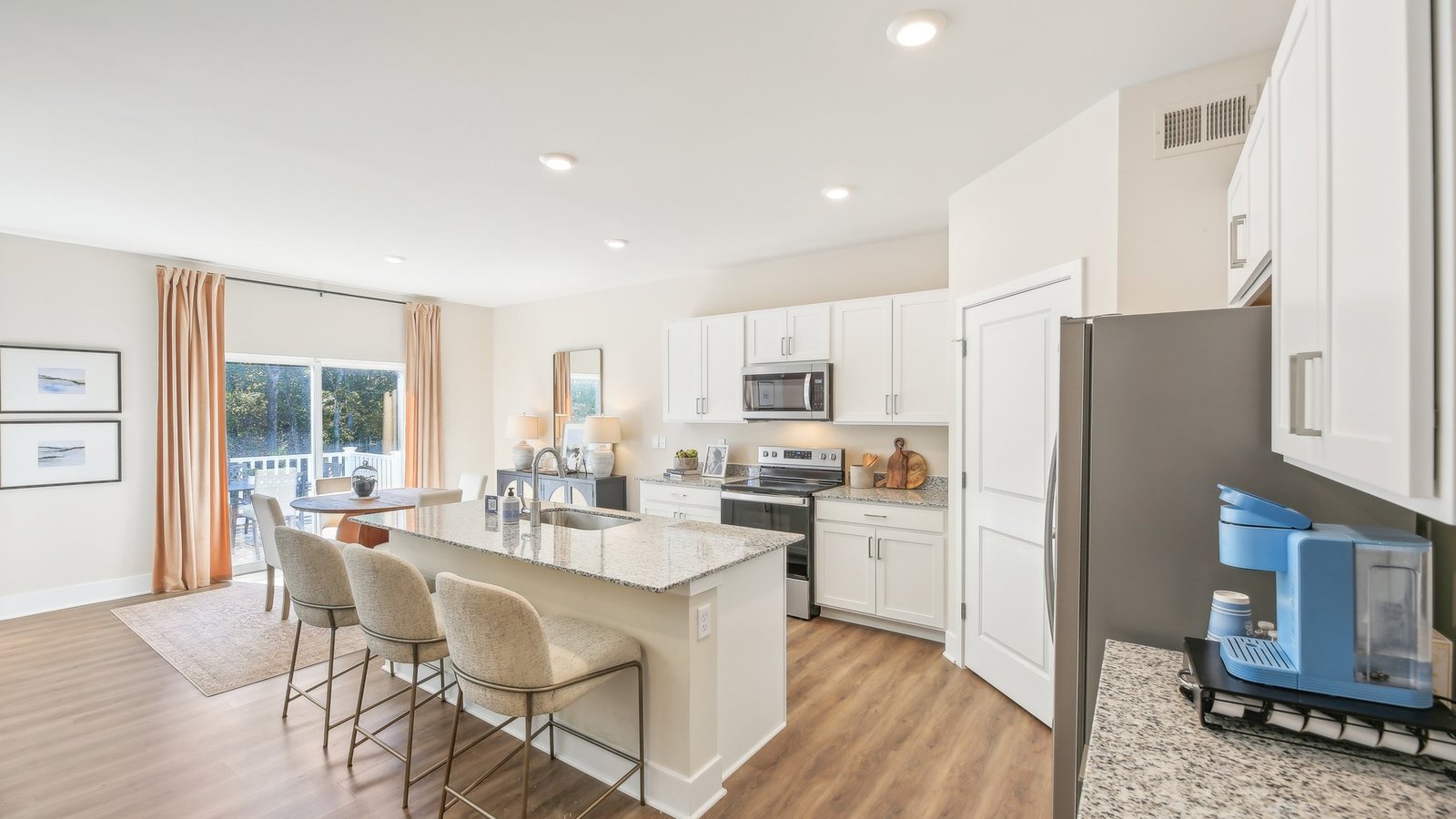 Modern kitchen and dining area with white cabinets, stainless steel appliances, a granite island with three chairs, wood flooring, and a window with curtains overlooking greenery.