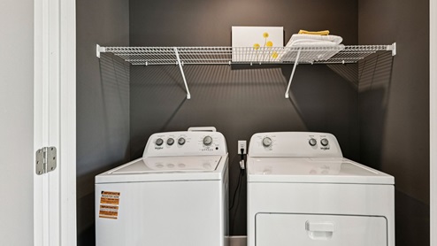 Laundry closet — side‑by‑side washer and dryer with wire shelf and decor.