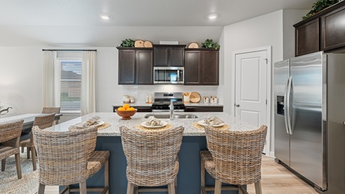A warm kitchen with dark shaker cabinets, a large center island with three woven barstools, and stainless-steel appliances.