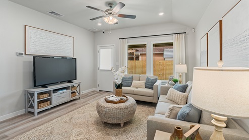 A living room with wood-look flooring, light couches, two windows with white curtains, a large tv, an entertainment system and a door to the backyard.