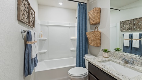 A bright bathroom with a dark vanity and light countertops, a toilet, a full shower and bathtub with a blue shower curtain and boho decor.