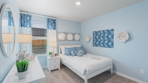 A baby blue bedroom with a neatly made bed, a large window with blue curtains, neutral carpets and a white bedside table.