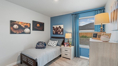A baseball-themed bedroom with one blue accent wall and neutral carpets, a twin bed, a white bedside table with a matching dresser, and a large window with blue striped curtains.