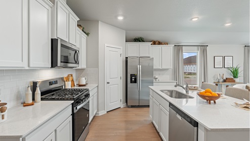 Side view of the kitchen area with white granite countertops, large island, white cabinets, white backsplash, and stainless steel appliances with pantry in the corner.