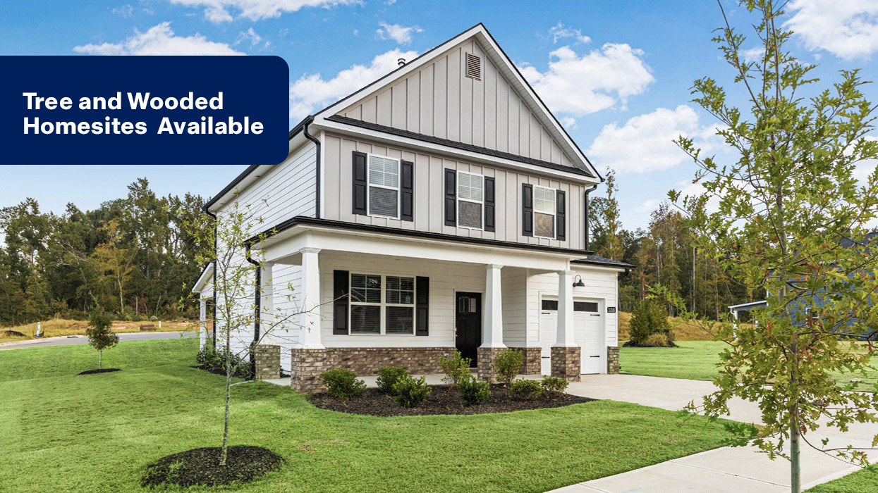 Two story white home with covered front porch and lush landscaping and banner saying "TREE AND WOODED HOMESITES AVAILABLE"