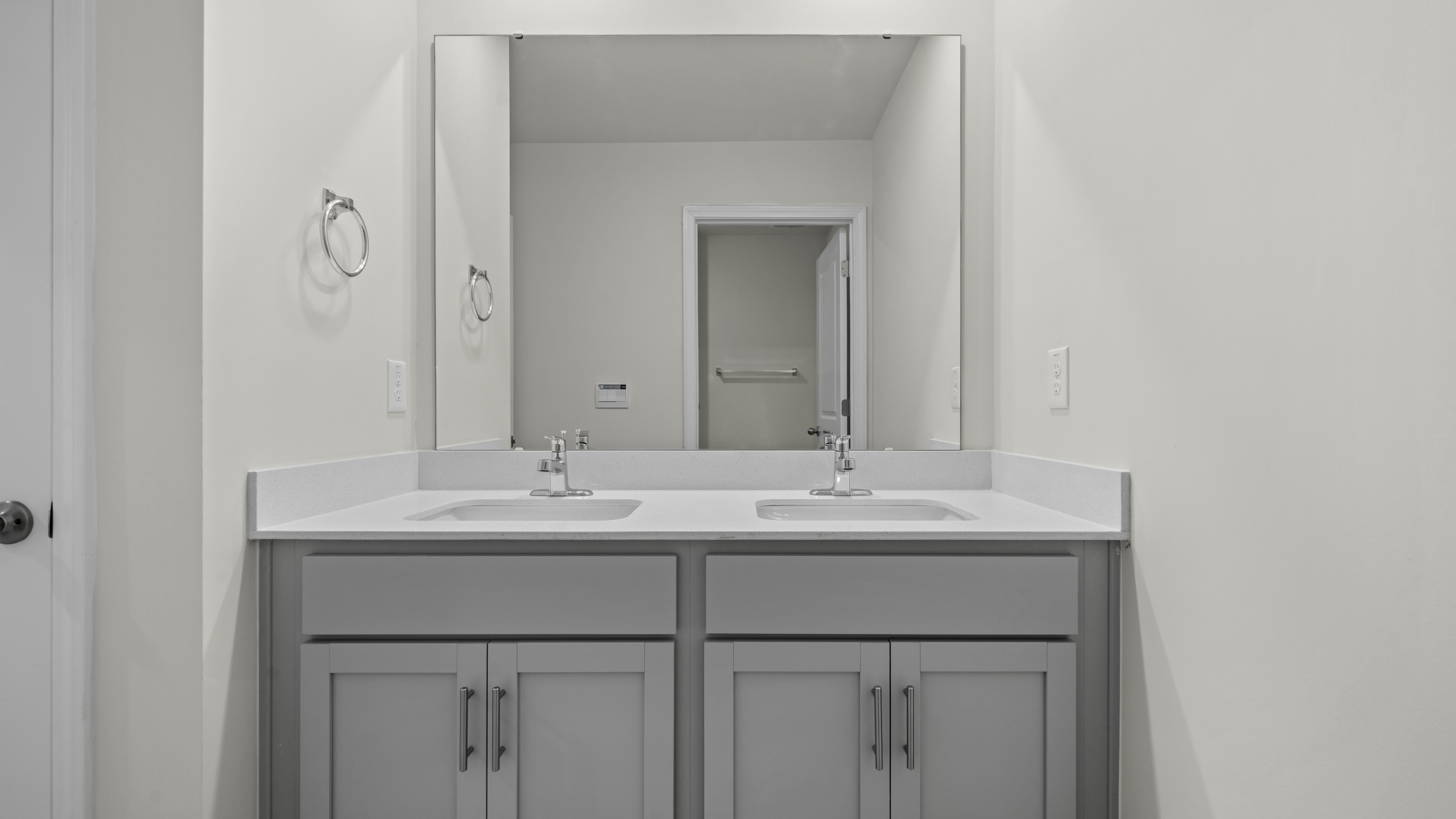Double vanity featuring light-gray shaker-style cabinetry, white solid-surface countertop, dual drop-in sinks, polished chrome faucets, and a large wall-mounted mirror