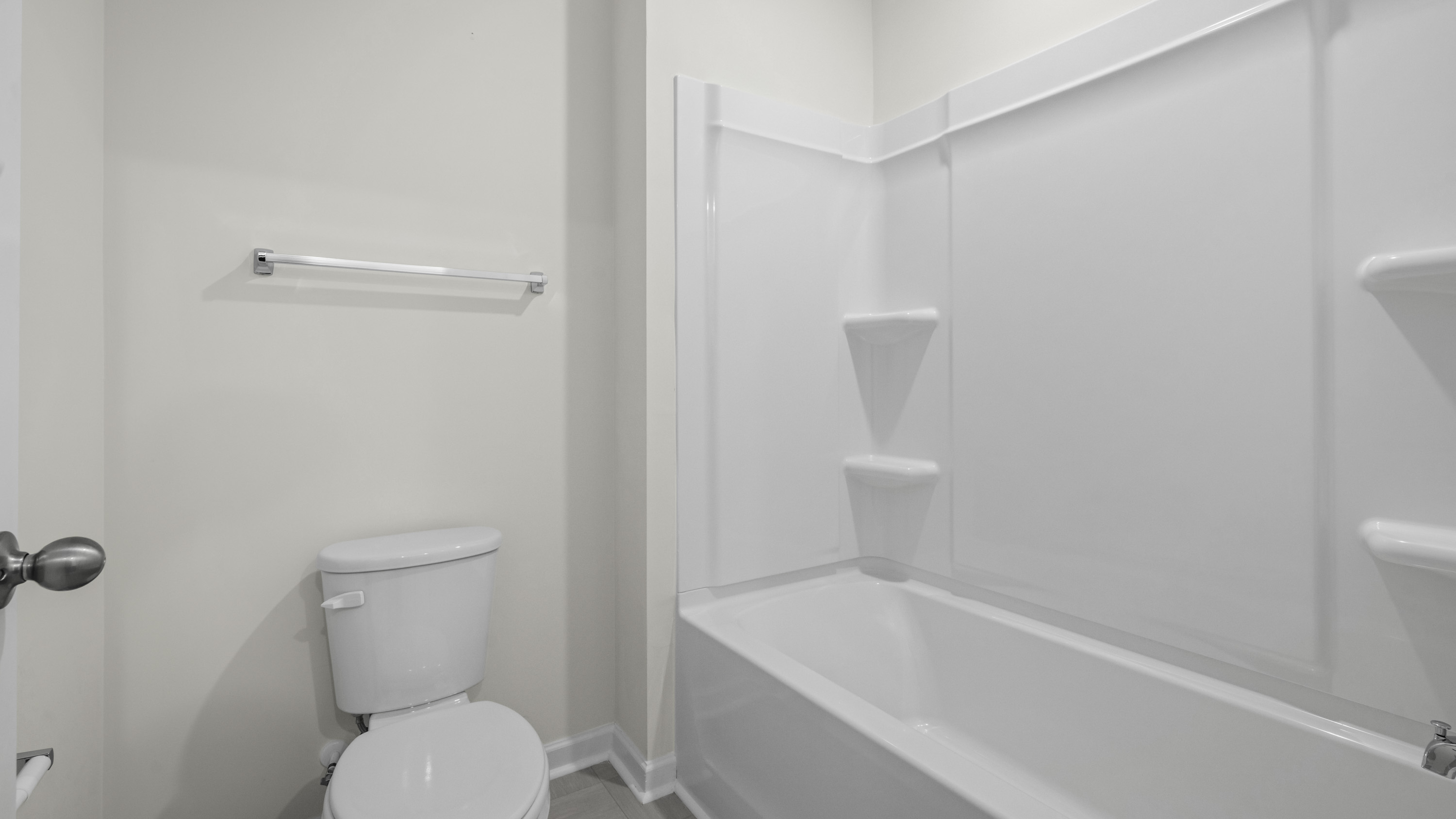 Bathroom featuring a white bathtub with integrated shower surround and shelving, dual-flush toilet, and wall-mounted metal towel bar