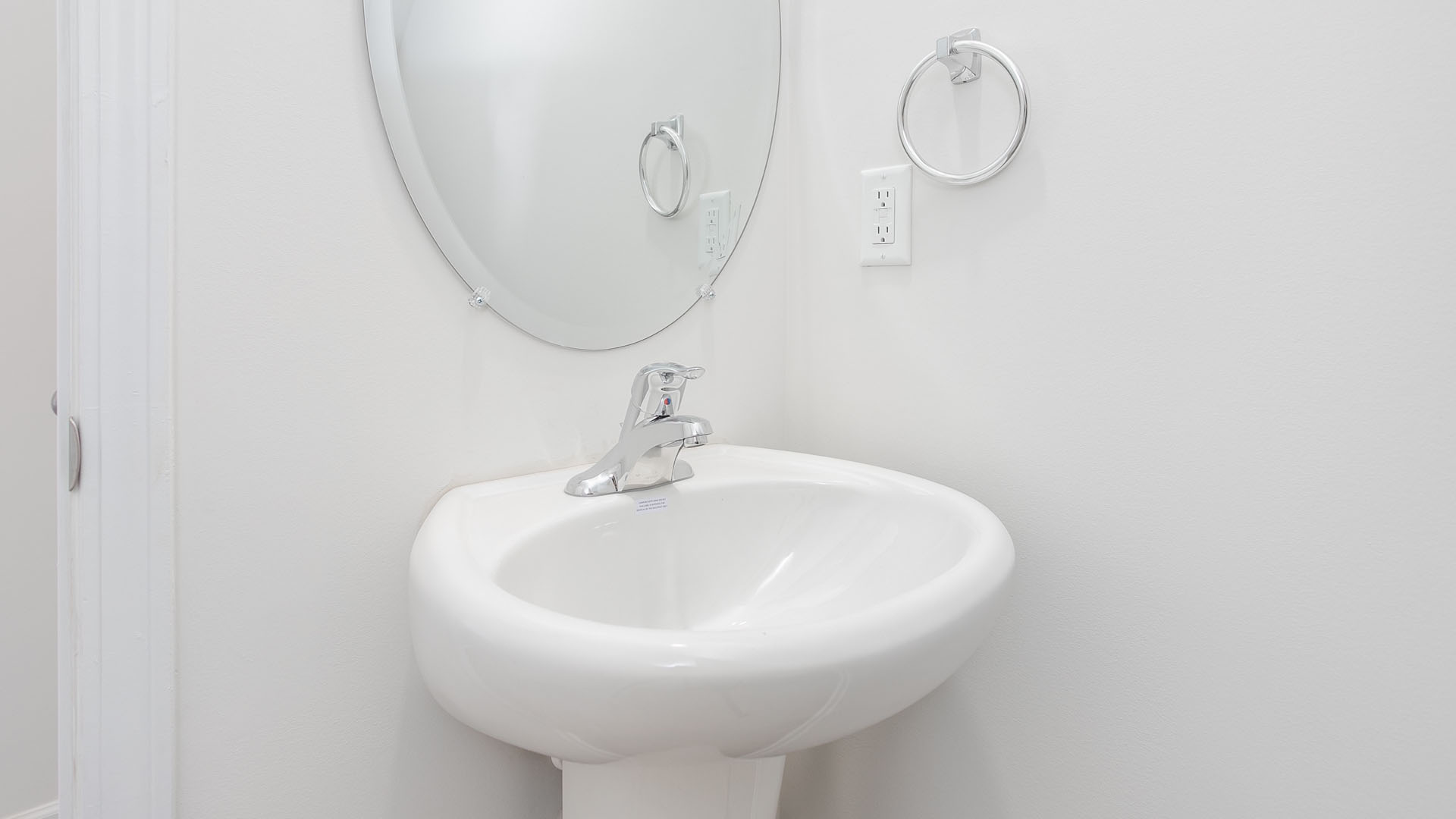 Wall-mounted oval mirror, white pedestal sink, polished chrome faucet, and chrome circular towel ring