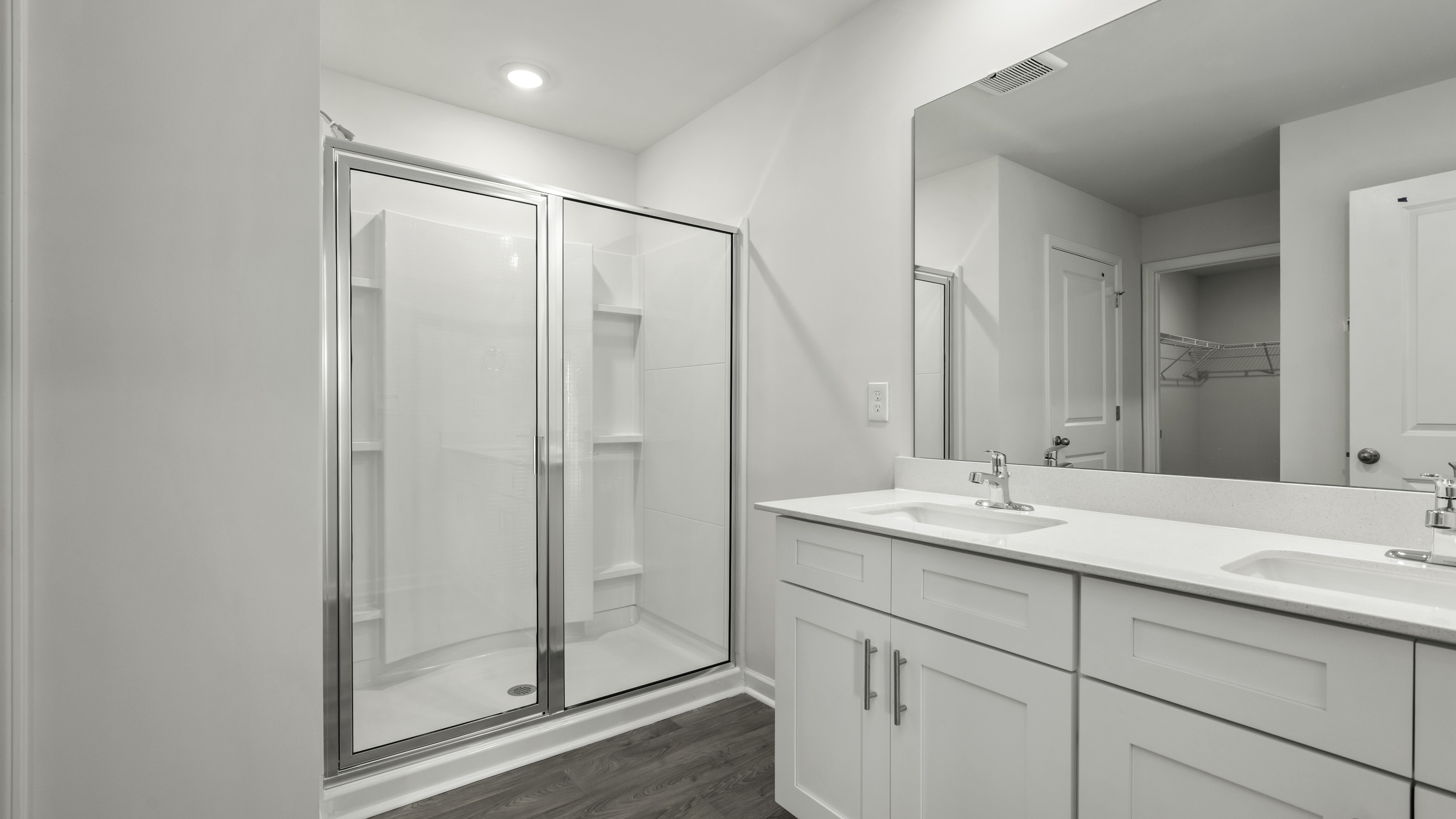 Double vanity with white cabinetry, solid surface countertops, chrome-finish fixtures, a large wall-mounted mirror, and a walk-in shower with a glass enclosure