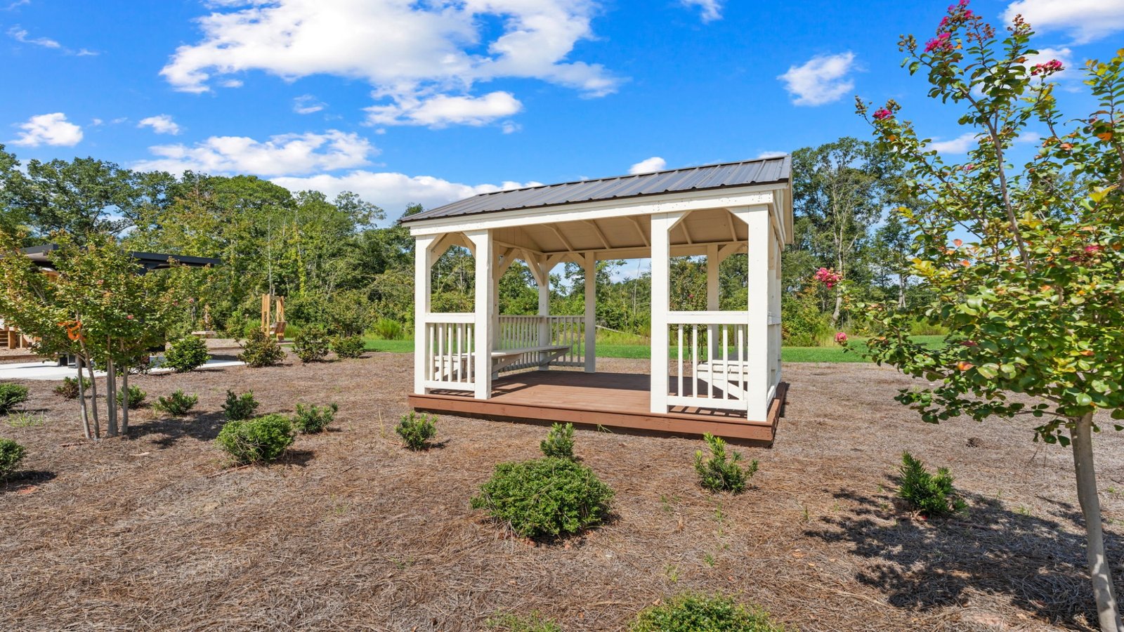 Community gazebo surrounded by landscaped grounds, flowering trees, and open green space under a bright blue sky.
