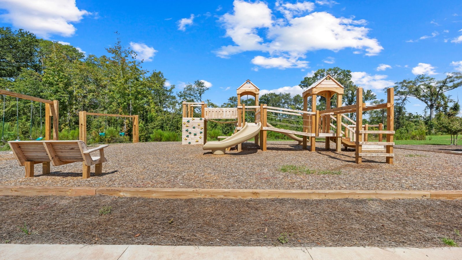 Community playground featuring swings, climbing wall, slides, and shaded seating area on a sunny day.
