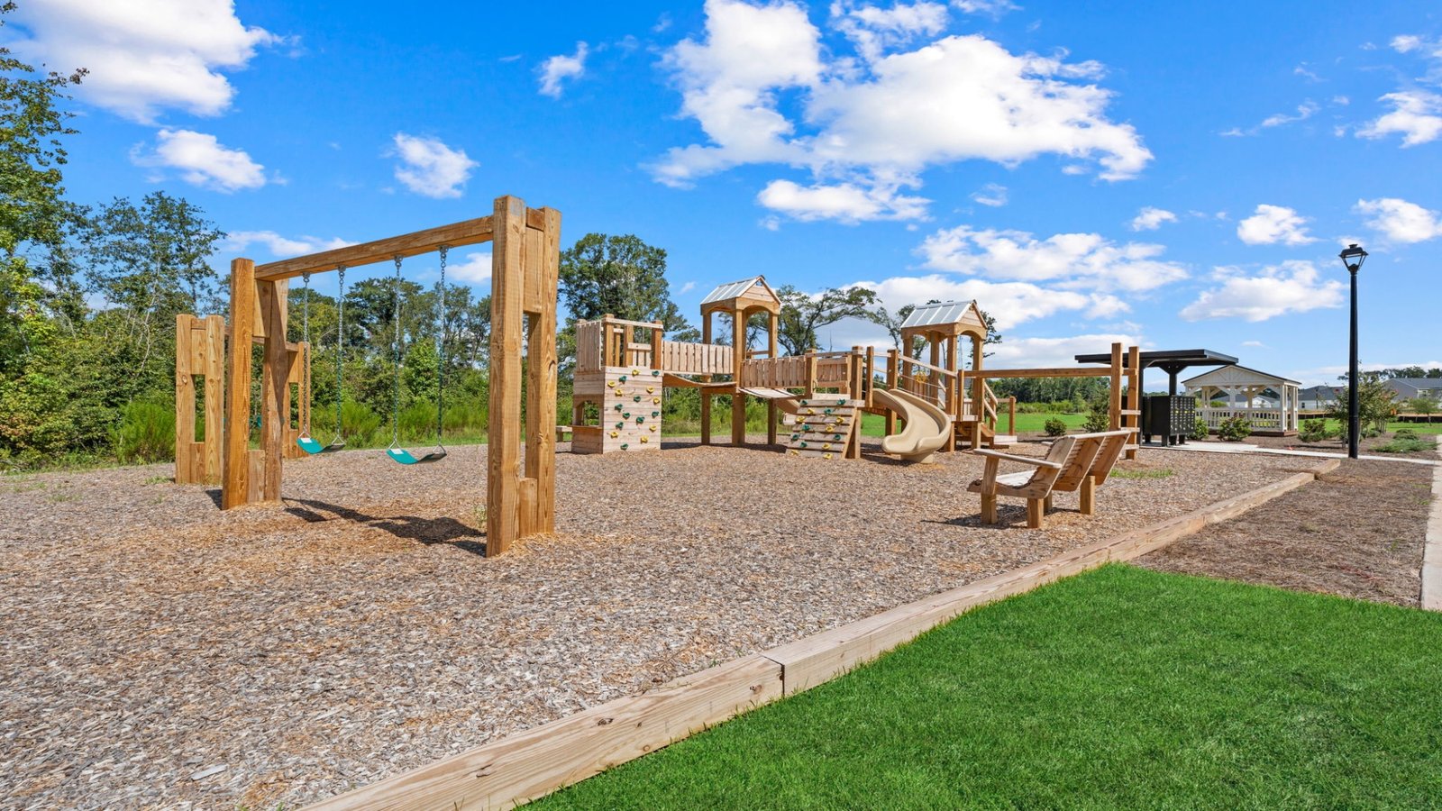 Neighborhood playground with wooden play structure, swings, and open green space surrounded by mature trees.