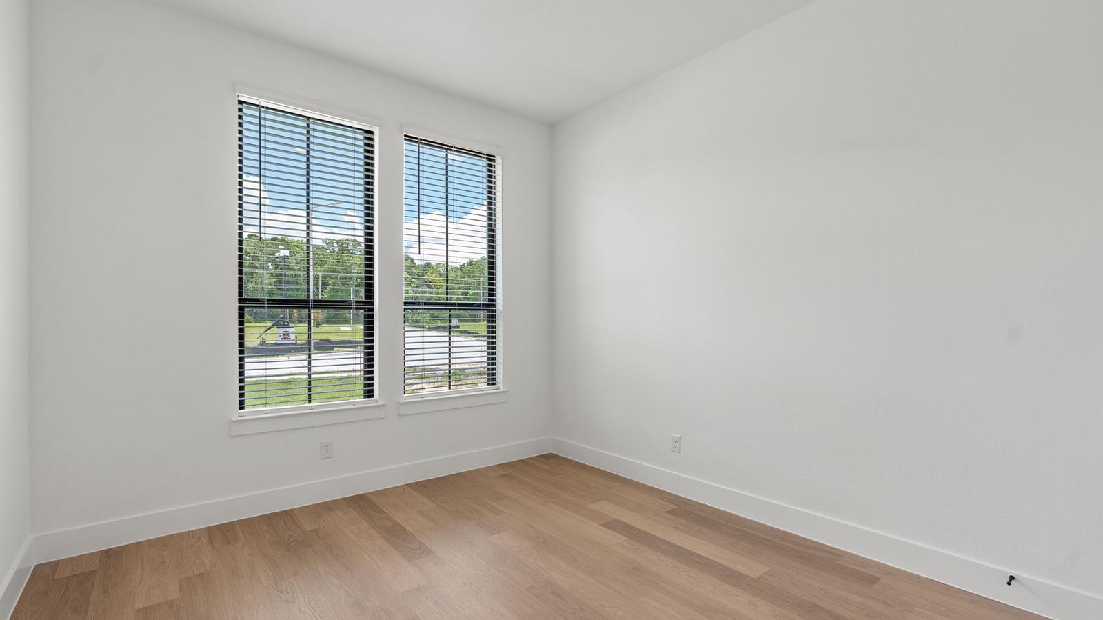 A secondary bedroom with two large windows, neutral painted walls, and wood flooring.