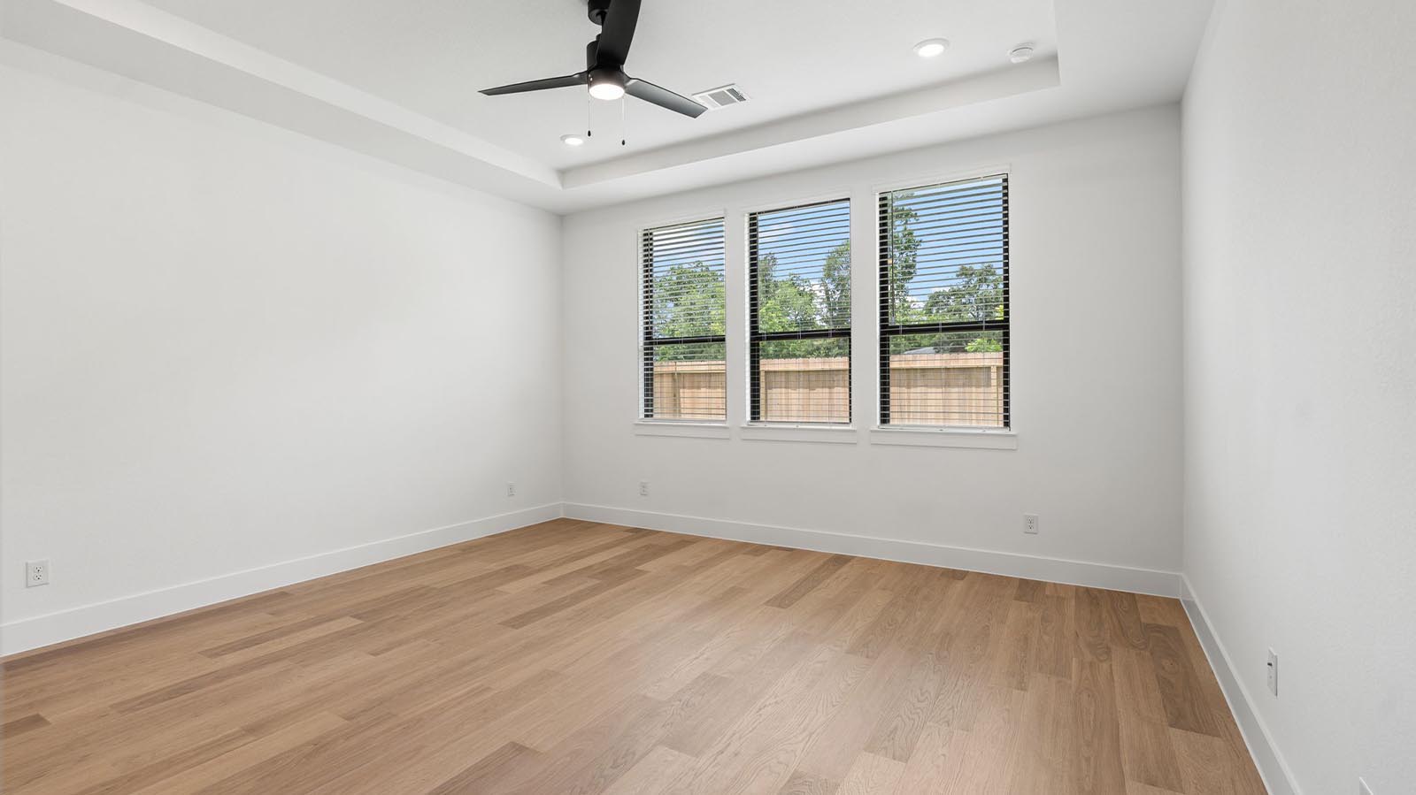 The primary bedroom with ceiling fan, three large windows for natural lighting, hardwood floors, and a large walk-in closet.