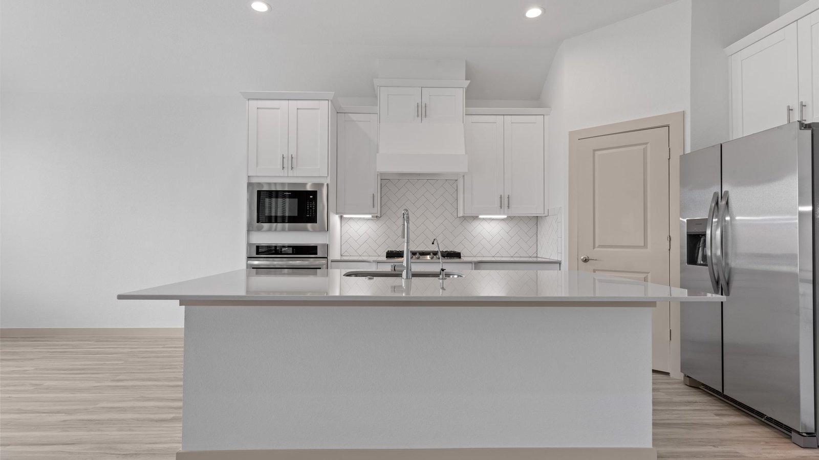 The kitchen features an island with a sink, light colored countertops, white shaker-style cabinets with under cabinet lighting, white subway tile backsplash in a herringbone pattern, stainless steel appliances and a pantry.