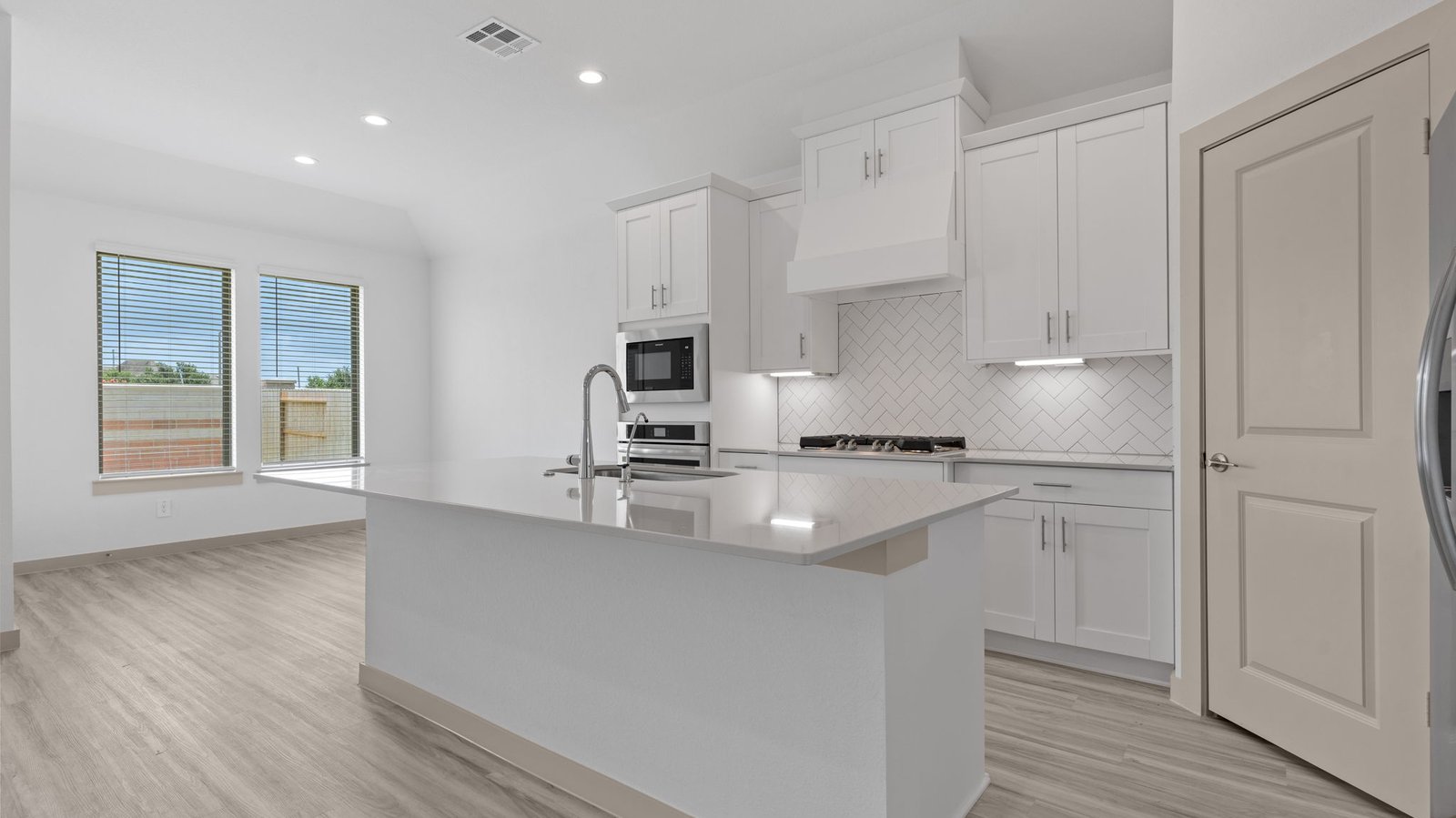 The kitchen features an island with a sink, light colored countertops, white shaker-style cabinets with under cabinet lighting, white subway tile backsplash in a herringbone pattern, stainless steel appliances, a pantry with a view of the breakfast/dining area with two windows.