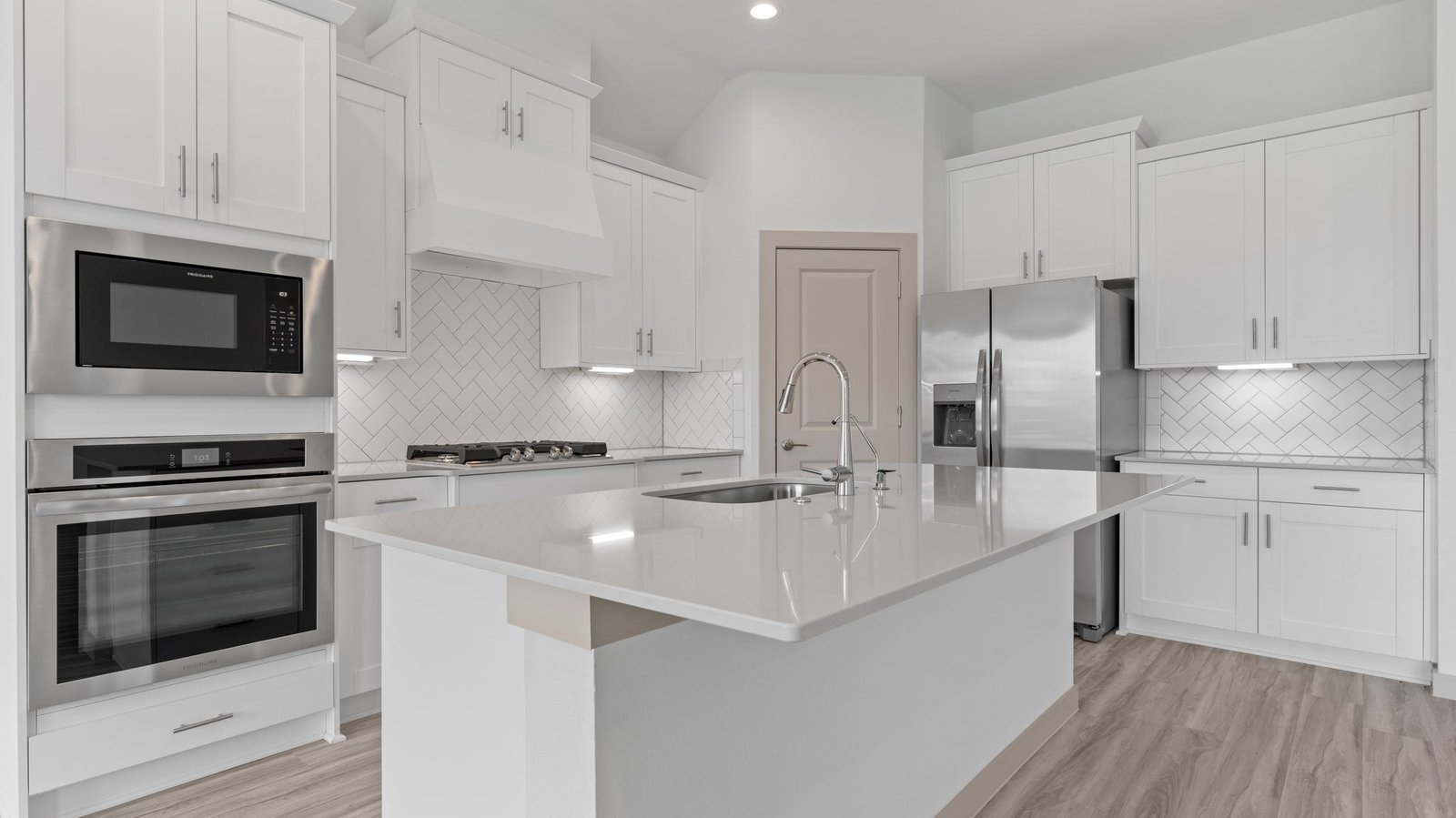 The kitchen features an island with a sink, light colored countertops, white shaker-style cabinets with under cabinet lighting, white subway tile backsplash in a herringbone pattern, stainless steel appliances and a pantry.