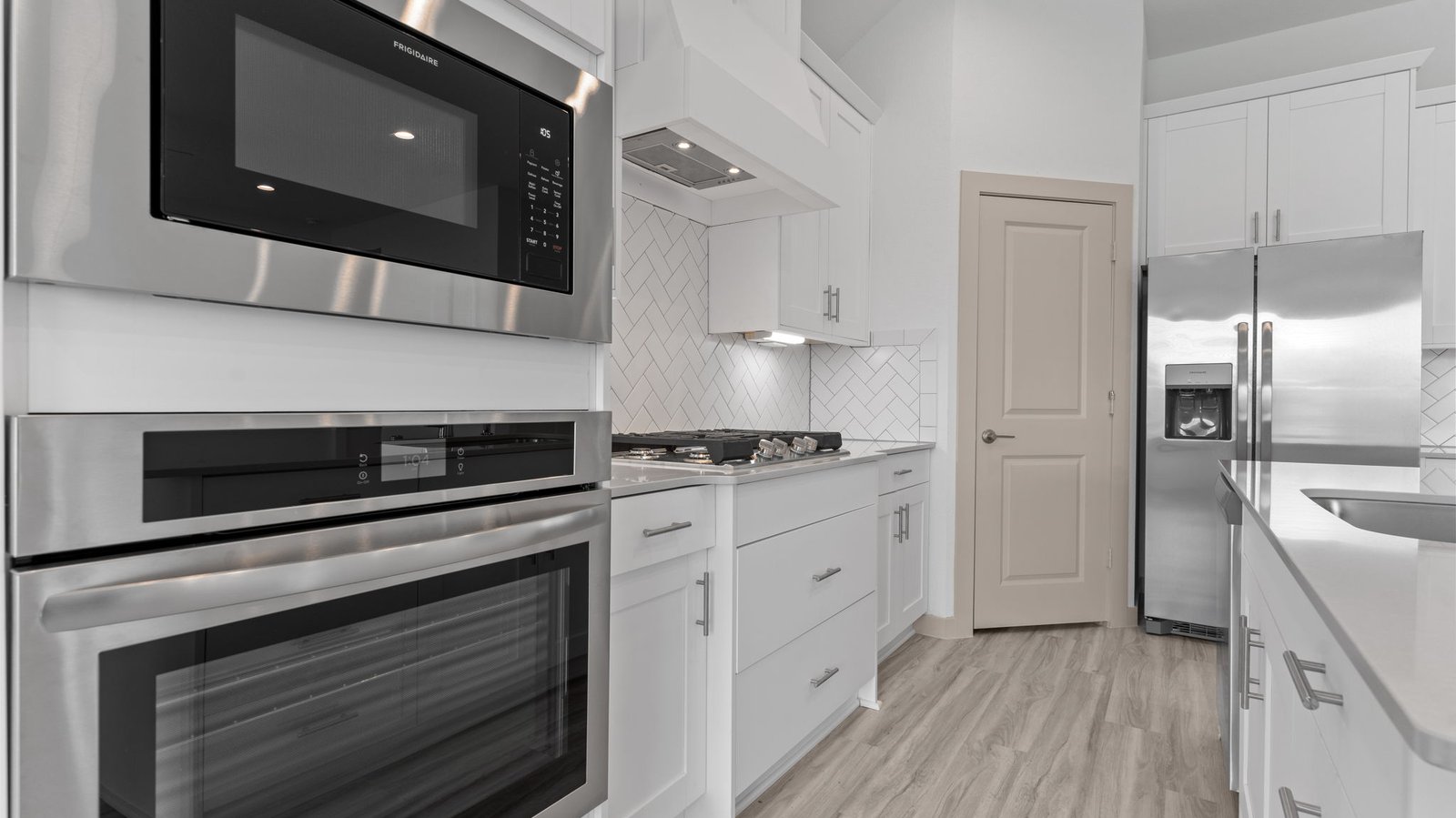 The kitchen features an island with a sink, light colored countertops, white shaker-style cabinets with under cabinet lighting, white subway tile backsplash in a herringbone pattern, stainless steel appliances and a pantry.