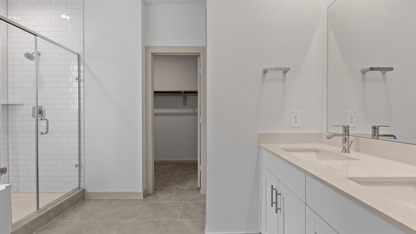 Master bathroom features a double vanity sink with light colored countertops, white-shaker style cabinets, a white subway tiled walk-in shower with a glass door and a view of the walk -in closet with neutral carpet.