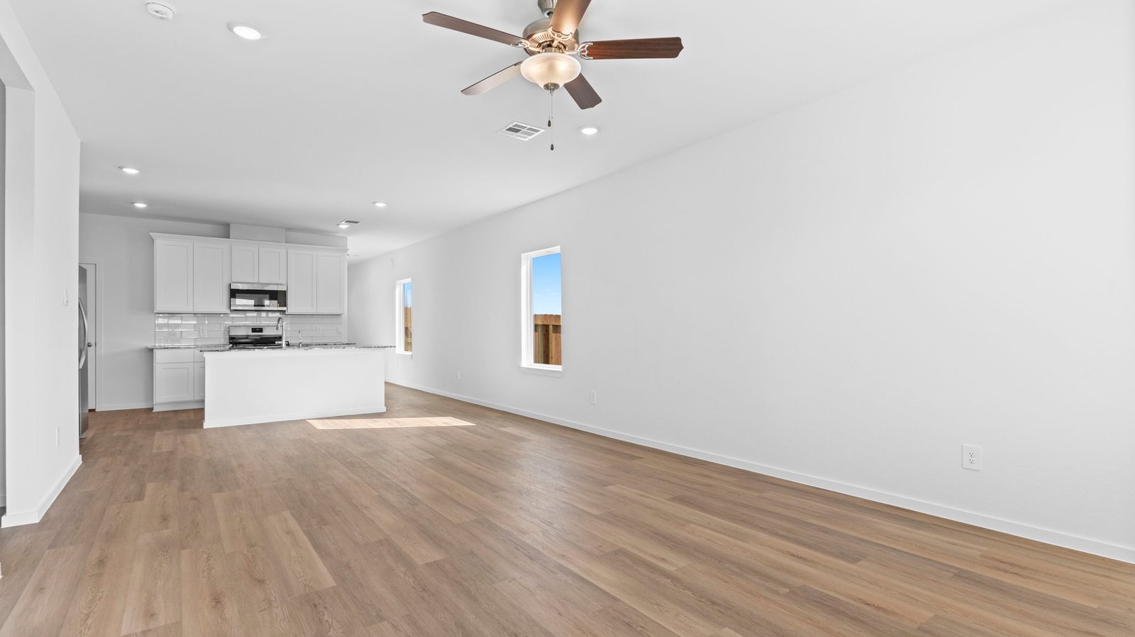 A cozy living room featuring large windows for natural lighting, vinyl wood flooring and a view to the kitchen and dining area.
