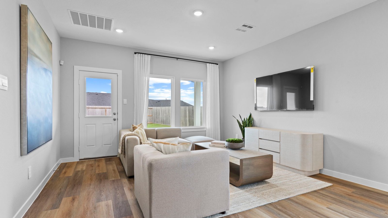 A cozy living room featuring large windows for natural lighting, wood flooring and a view to the kitchen and dining area.