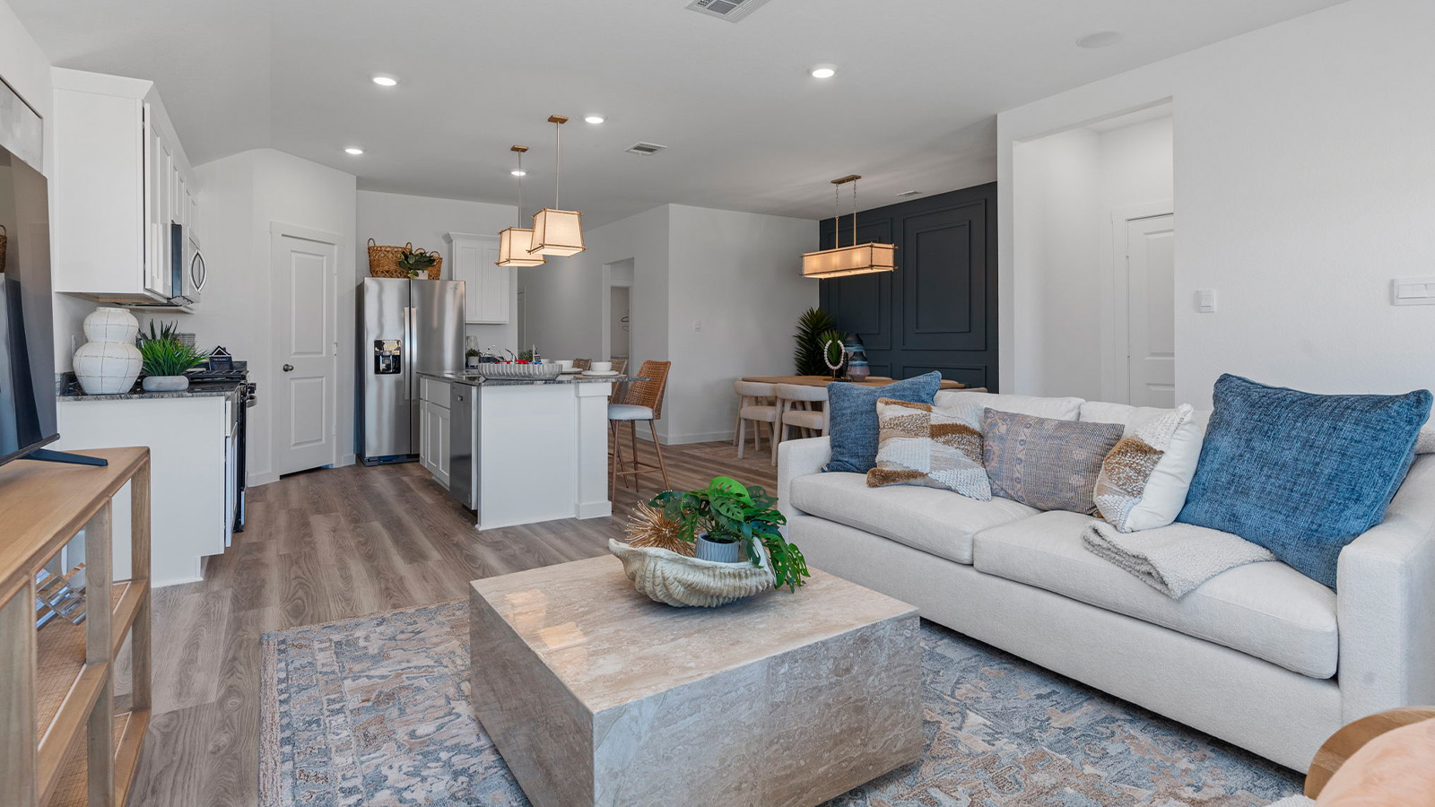 A cozy living room featuring large windows for natural lighting, wood flooring and a view to the kitchen and dining area.